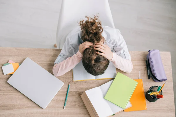 Top view of schoolchild holding hands on head while sitting at wooden desk and doing schoolwork at home — Stock Photo