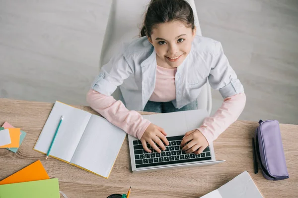 Vista aérea del niño adorable usando el ordenador portátil mientras está sentado en el escritorio de madera y haciendo el trabajo escolar en casa - foto de stock