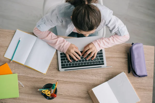 Top view of schoolchild using laptop while sitting at desk near copy books and stationery — Stock Photo