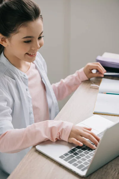 Écolier joyeux en utilisant un ordinateur portable tout en étant assis au bureau en bois et faire des devoirs — Stock Photo