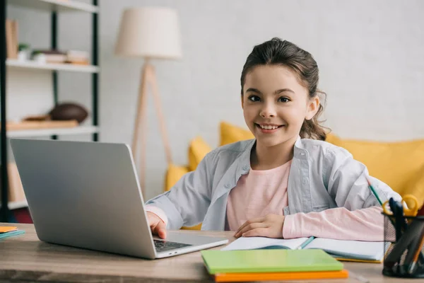 Щаслива дитина дивиться на камеру, сидячи за столом і використовуючи ноутбук вдома — стокове фото