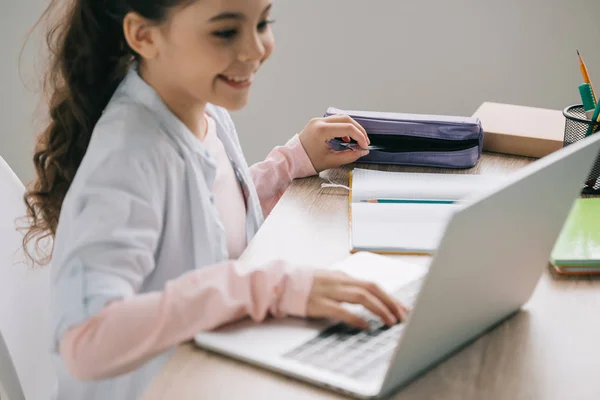 Selective focus of smiling schoolchild getting ruler out of pencil case while using laptop at home — Stock Photo