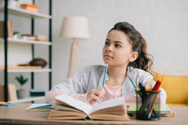 Nachdenkliches Kind hält Bleistift in der Hand und schaut weg, während es zu Hause Hausaufgaben macht — Stockfoto