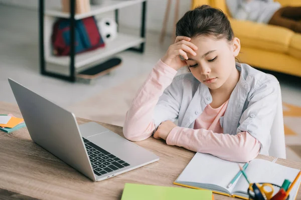 Exhausted schoolchild sitting at desk near laptop and copy book while doing schoolwork at home — Stock Photo