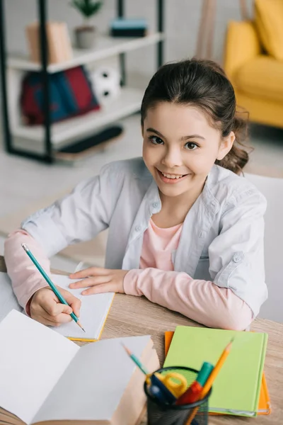 Alegre colegial escribiendo en el cuaderno y sonriendo a la cámara mientras hace la tarea - foto de stock