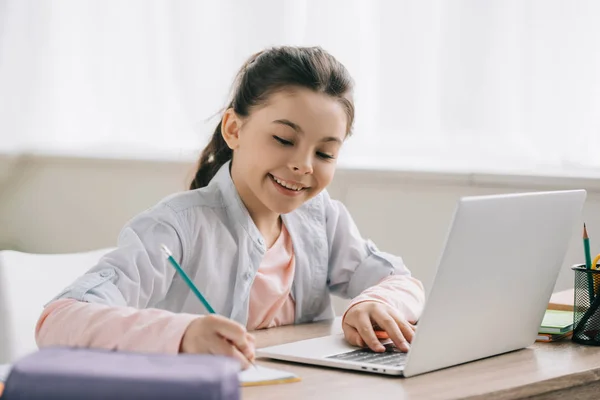 Очаровательный улыбающийся ребенок пишет в копировальной книге и с помощью ноутбука во время выполнения домашних заданий — стоковое фото