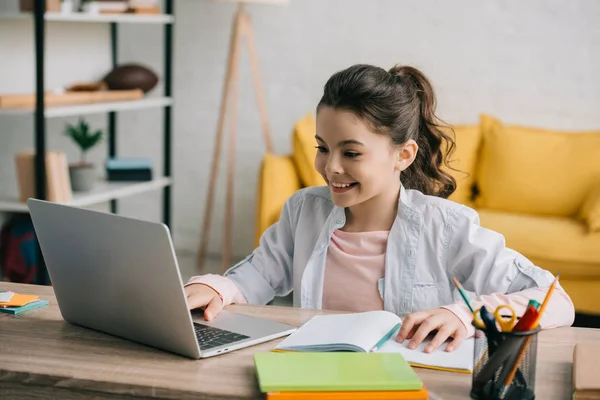 Mignon enfant souriant en utilisant un ordinateur portable tout en étant assis au bureau avec des livres de copie et de faire des devoirs — Photo de stock