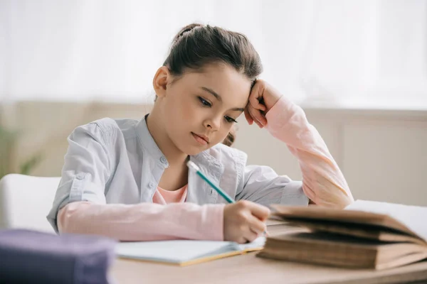 Nachdenkliches Schulkind schreibt in Notizbuch, während es am Schreibtisch sitzt und Hausaufgaben macht — Stockfoto