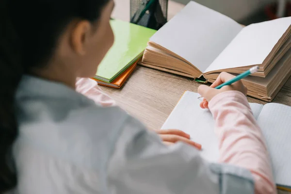 Вид ззаду шкільної дитини, що пише в блокноті, сидячи на столі і роблячи домашнє завдання — стокове фото