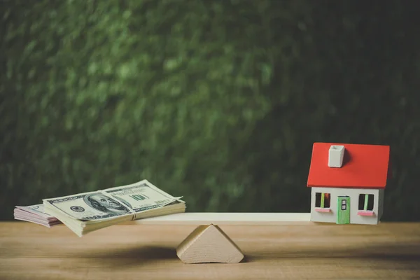 Dollar banknotes and house model balancing on seesaw on green background — Stock Photo