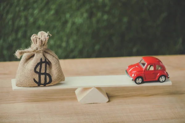 Червоний мініатюрний автомобіль і грошовий мішок з доларовим знаком балансування на базі на зеленому фоні — стокове фото