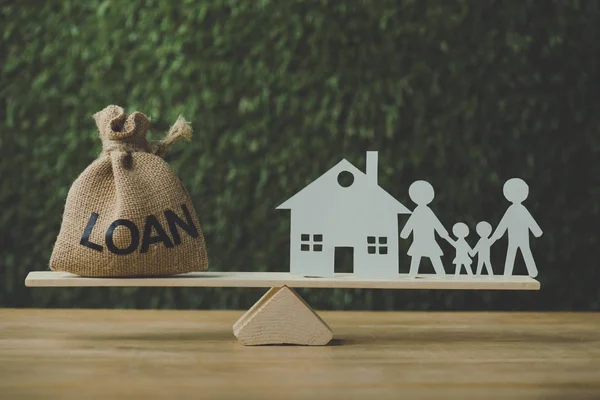 Paper house and paper cut family balancing on swing with money bag with loan inscription on green background — Stock Photo