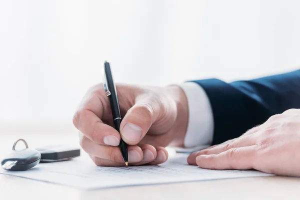 Vista parcial del empresario sosteniendo la pluma y la escritura en el acuerdo de préstamo y las llaves del coche en la mesa — Stock Photo