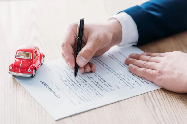 Vue partielle du concessionnaire automobile écrivant dans l'accord de prêt près de voiture miniature rouge sur table en bois — Photo de stock