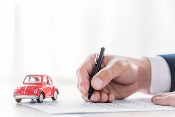 Vue partielle du concessionnaire automobile écrivant dans l'accord de prêt près de voiture miniature rouge sur la table — Photo de stock