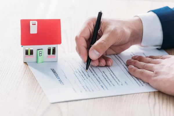 Vue partielle de l'écriture d'agent immobilier dans l'accord de prêt près du modèle de maison sur le dessus de table en bois — Photo de stock