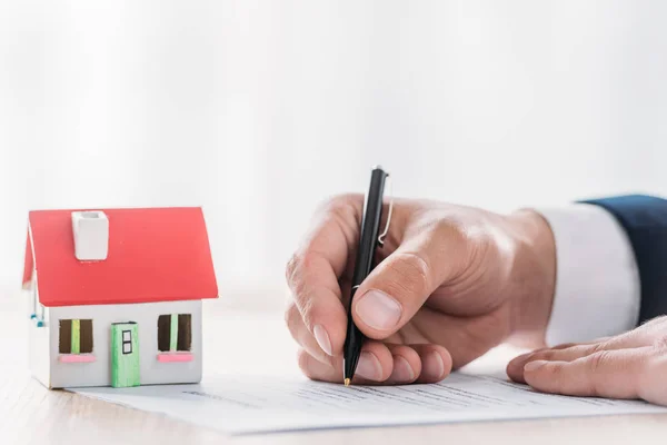 Vista recortada de escritura de bienes raíces en contrato de préstamo cerca de modelo de casa en la mesa - foto de stock