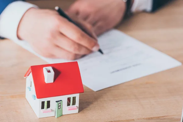 Vista recortada de hombre de negocios escribiendo en contrato de préstamo cerca de modelo de casa en la mesa de madera - foto de stock