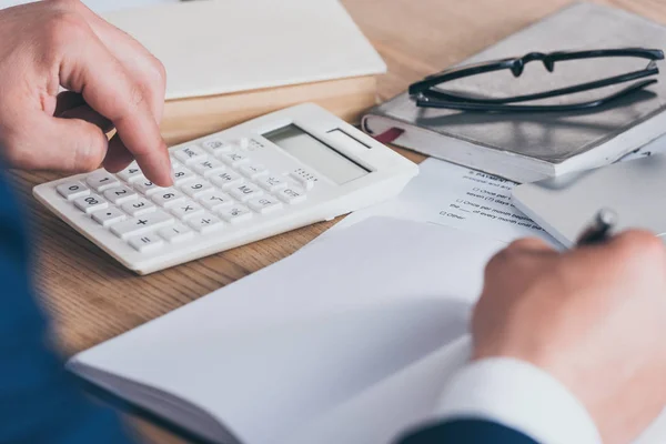 Vista parcial del gerente escribiendo en el cuaderno y utilizando la calculadora en el lugar de trabajo - foto de stock