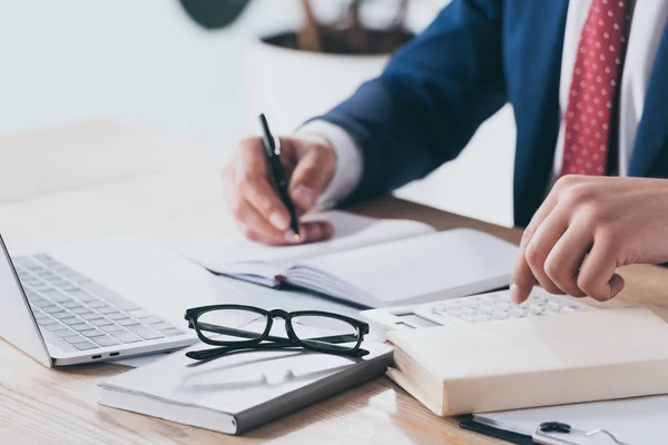 Vista parcial del hombre de negocios en ropa formal usando calculadora y escritura en cuaderno en el lugar de trabajo - foto de stock