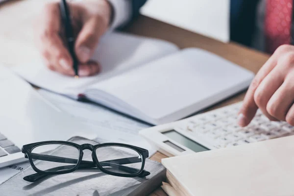 Recortado tiro de hombre de negocios escribir en cuaderno vacío mientras se utiliza la calculadora - foto de stock