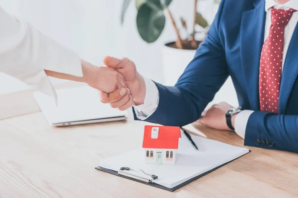 Partial view of businessman and client shaking hands near house model on table — Stock Photo