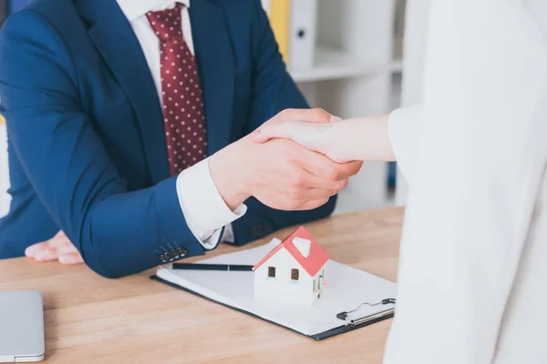 Vista parcial del cliente estrechando la mano con el agente inmobiliario cerca del modelo de la casa — Stock Photo