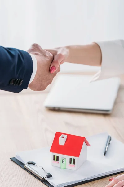 Vista parcial del hombre de negocios estrechando la mano con el cliente cerca del modelo de la casa en la mesa — Stock Photo