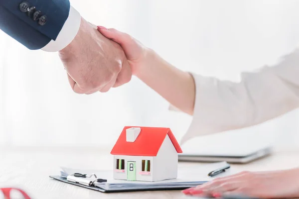 Cropped shot of realtor shaking hands with client near house model on tabletop — Stock Photo