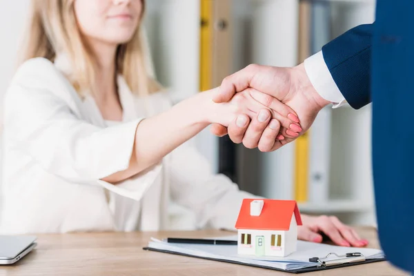 Vista parcial de la mujer sonriente estrechando la mano con el modelo de la casa cerca de la casa en la mesa - foto de stock