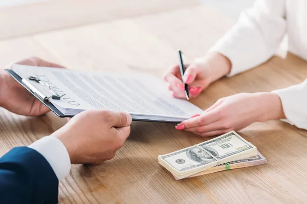 Vista parcial de la mujer firmando contrato en el portapapeles en manos de un hombre de negocios cerca de billetes de dólar en la mesa - foto de stock