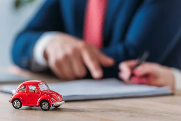 Tiro recortado de la pluma de retención del cliente y concesionario de automóviles apuntando al lugar de firma en el contrato cerca de coche de juguete rojo en la mesa - foto de stock