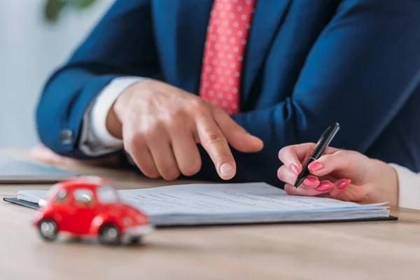 Partial view of client holding pen and car dealer pointing at signature place in loan agreement near red toy car on table — Stock Photo