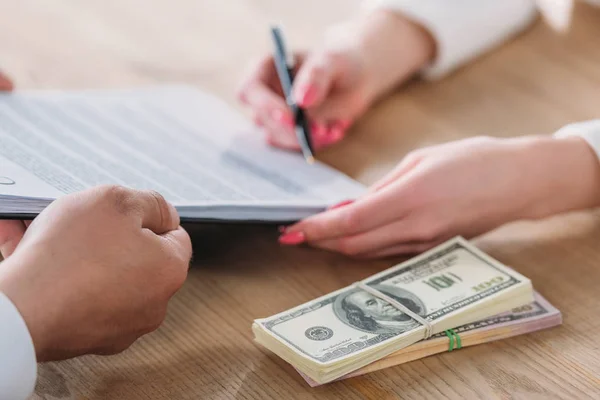 Vista recortada de la mujer firmando un acuerdo de préstamo en el portapapeles en manos de hombres de negocios cerca de billetes de dólar - foto de stock