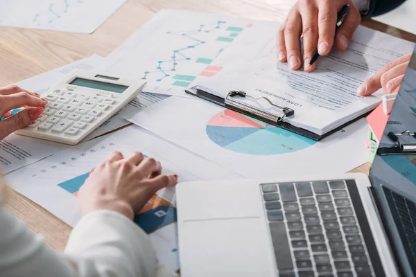 Visión parcial del hombre que estudia el acuerdo de préstamo y la mujer de negocios usando calculadora mientras está sentado en el lugar de trabajo con gráficos y gráficos - foto de stock