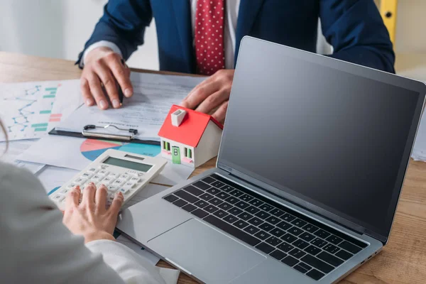 Recortado tiro de hombre estudiando acuerdo de préstamo y mujer de negocios usando calculadora mientras está sentado en el lugar de trabajo con cerca de la computadora portátil, modelo de casa, gráficos y gráficos - foto de stock
