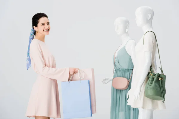 Улыбающаяся молодая женщина с пакетами для покупок возле манекенов, изолированных на сером — стоковое фото
