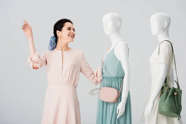 Bela menina sorridente gesticulando perto de manequins em vestidos isolados em cinza — Fotografia de Stock