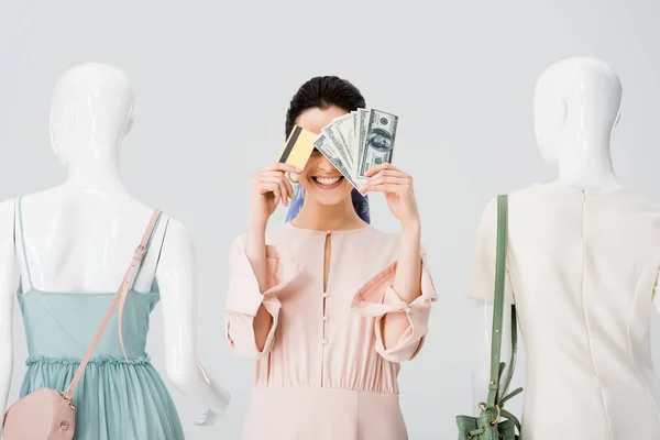 Hermosa chica cerca de maniquíes cubriendo la cara con dinero y tarjeta de crédito aislado en gris - foto de stock