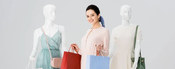 Panoramic shot of beautiful young woman with shopping bags near mannequins isolated on grey — Stock Photo