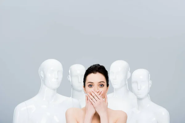Beautiful young woman covering mouth with hands while posing near mannequins isolated on grey — Stock Photo