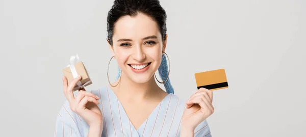 Plano panorámico de hermosa chica elegante sosteniendo caja de regalo y tarjeta de crédito aislada en gris - foto de stock