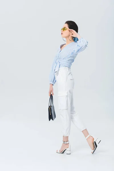 Красива стильна молода жінка в сонцезахисних окулярах тримає сумку і позує на сірому — Stock Photo