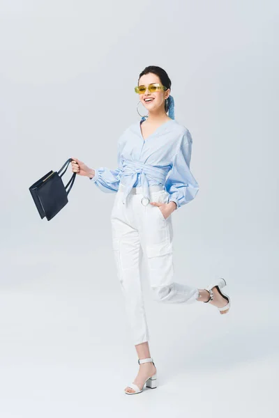 Belle jeune femme élégante dans des lunettes de soleil tenant sac et souriant sur gris — Photo de stock