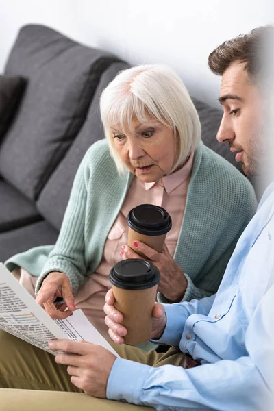 Homem e surpreendido mãe sênior segurando café e ler jornal em casa — Fotografia de Stock