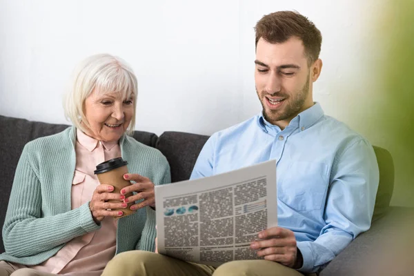 Улыбающаяся старшая мать держит кофе и читает газету с сыном дома — стоковое фото