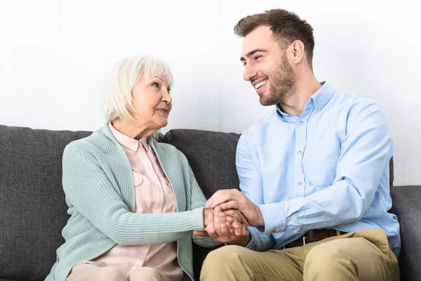 Souriant mère aînée et fils tenant la main et se regardant tout en étant assis sur le canapé — Photo de stock