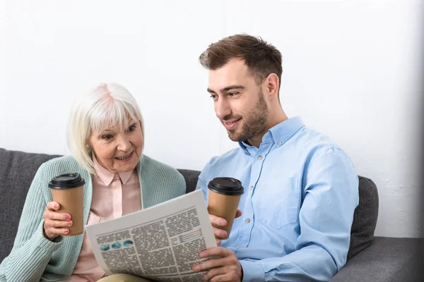 Homme souriant et surpris mère aînée tenant du café et lire le journal à la maison — Photo de stock
