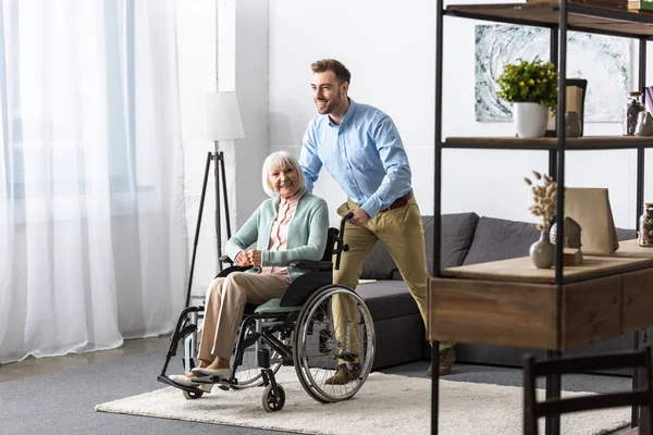 Lächelnder Mann trägt behinderte Seniorin im Rollstuhl — Stockfoto