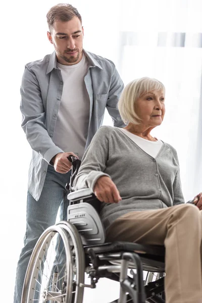 Bearded man carrying disabled mother on wheelchair at home — Stock Photo